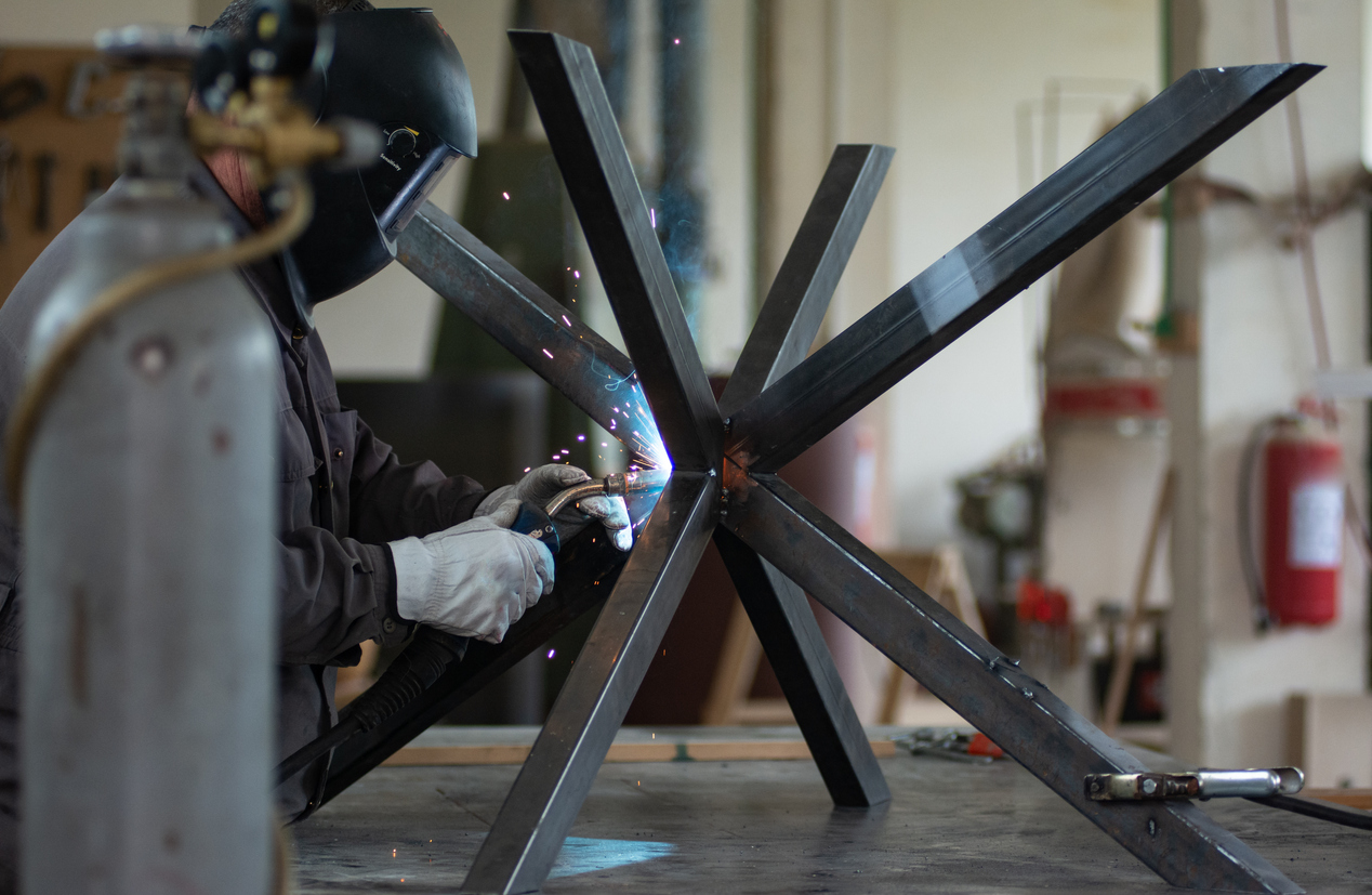 A mature blacksmith welds the metal structure of a dining table in a small workshop