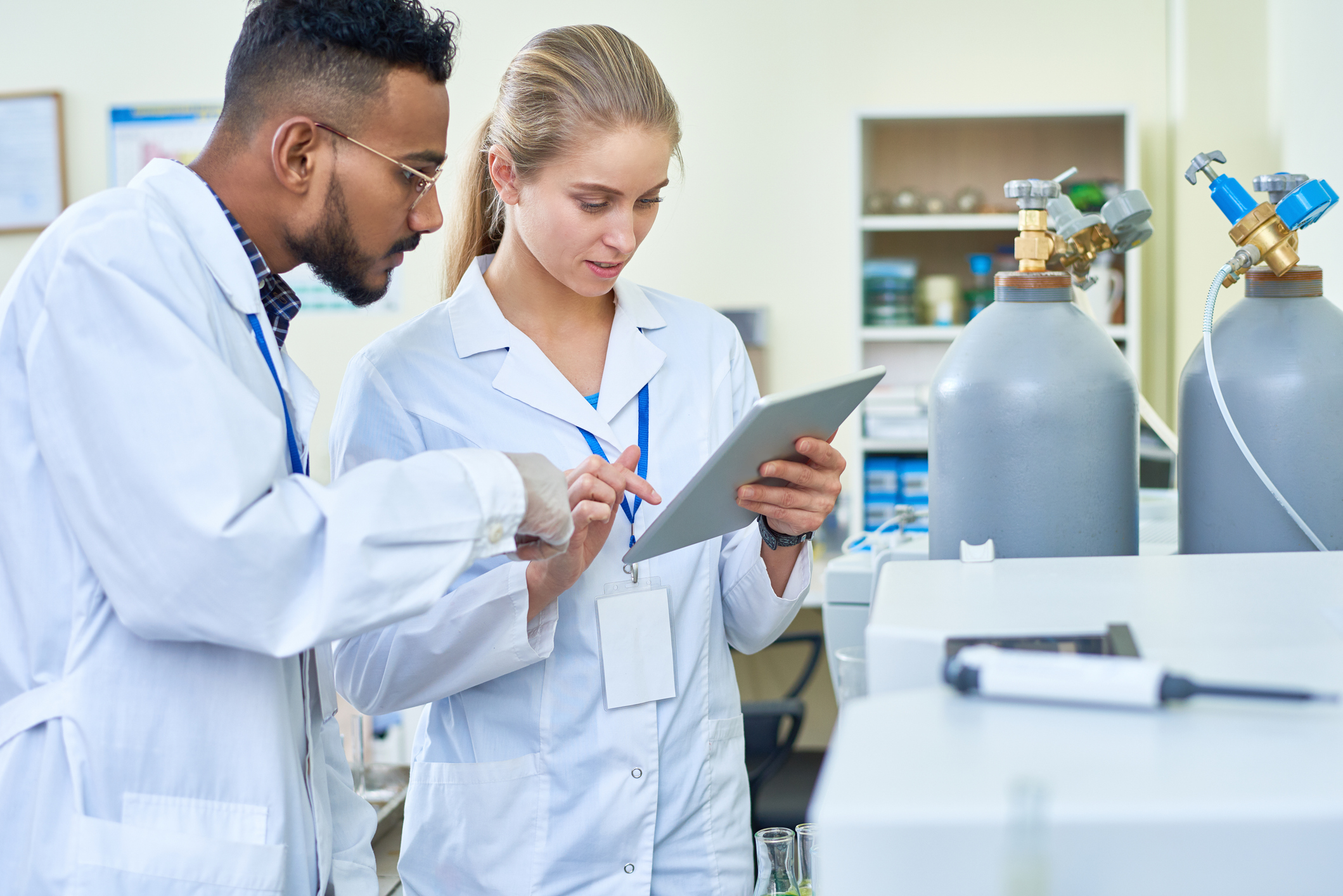 Serious thoughtful medical colleagues using digital tablet while viewing report together and discussing results standing by oxygen cylinders in laboratory